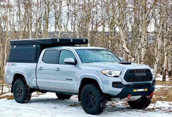Ein grauer Pickup-Truck mit Überdachung steht in einer verschneiten Waldlandschaft.