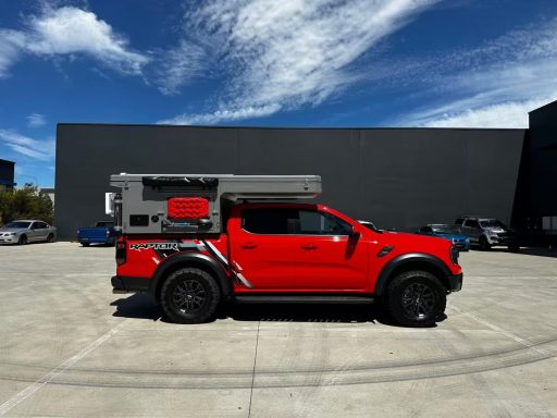 Roter Pickup mit Camperaufbau, geparkt vor einem grauen Gebäude unter blauem Himmel.