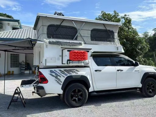 Weißer Pickup mit Wohnaufbau, parkend auf einem Kiesplatz, umgeben von Bäumen.